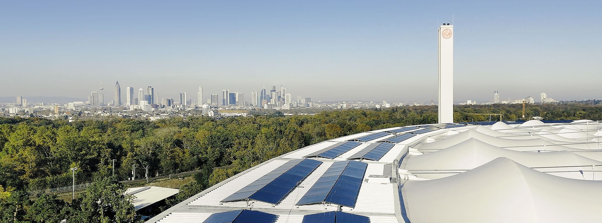 Solarmodule auf einem geschwungenen Dach, dahinter Frankfurts Skyline und viel Grün bei klarem Himmel im Panorama.