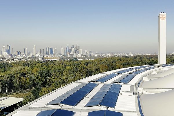 Solarmodule auf einem geschwungenen Dach, dahinter Frankfurts Skyline und viel Grün bei klarem Himmel im Panorama.