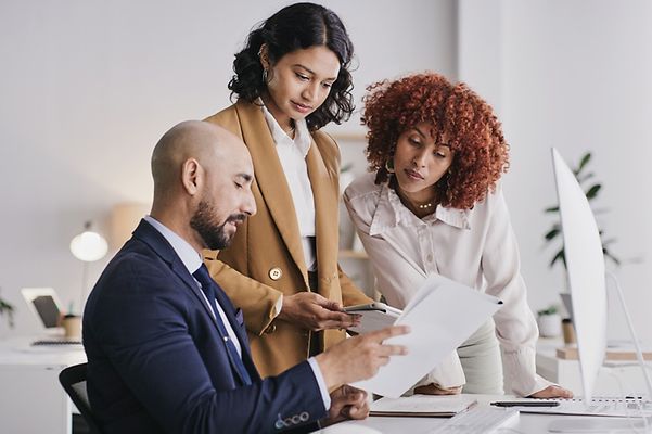 Ein Mann sitzt am Schreibtisch und hat Papiere vor sich, zwei Frauen beugen sich darüber