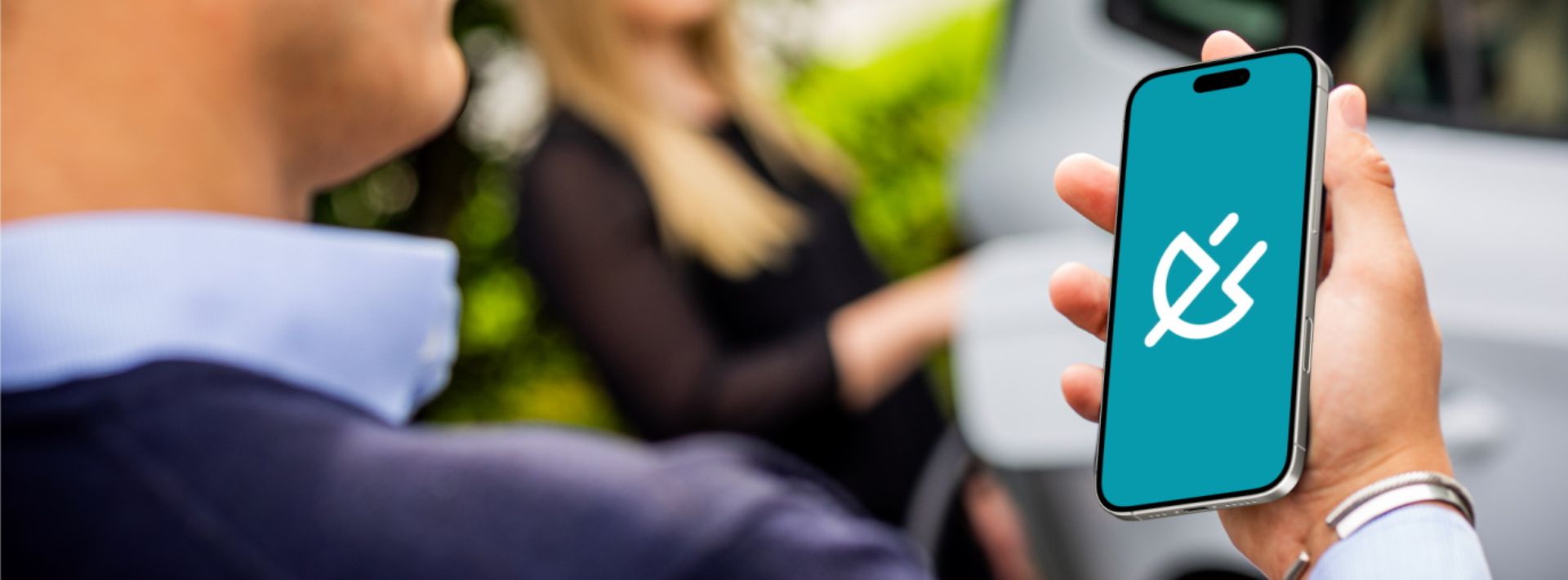 Eine Person hält ein Smartphone, trägt ein dunkelblaues Oberteil. Im Hintergrund steht eine Person vor einem weißen Auto.