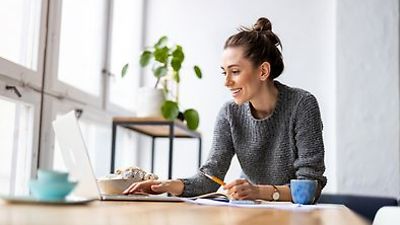 Frau sitzt vor einem Laptop, mit Stift und Block