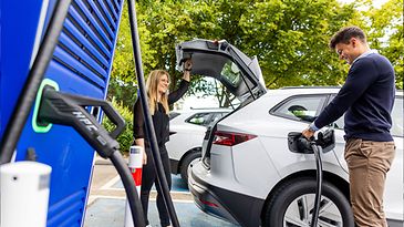 Frau und Mann mit weißem E-Auto an Ladesäule, Mann lädt Auto