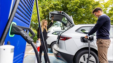 Frau und Mann mit weißem E-Auto an Ladesäule, Mann lädt Auto