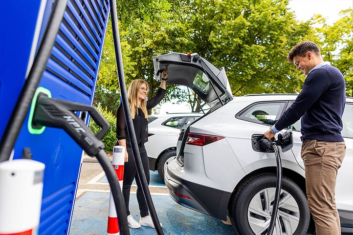 Frau und Mann mit weißem E-Auto an Ladesäule, Mann lädt Auto