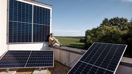 Junger Mann mit Arbeitshandschuhen sitzt auf einem Balkon mit drei großen Solarpanelen