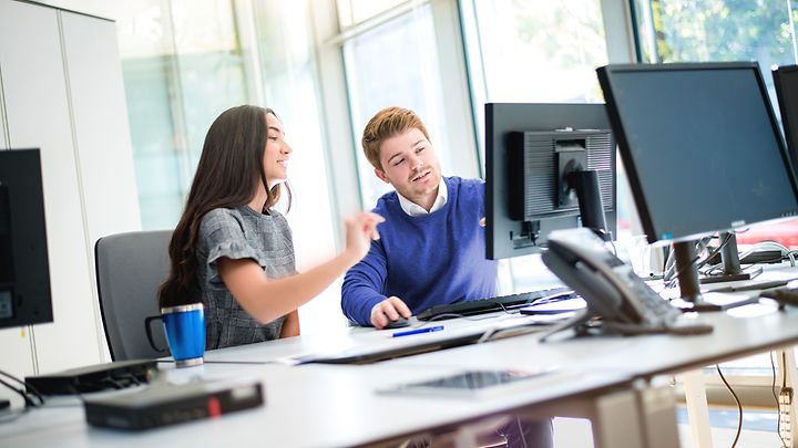 Zwei Personen sitzen im Büro vor Monitoren, eine zeigt erklärend auf den Bildschirm.
