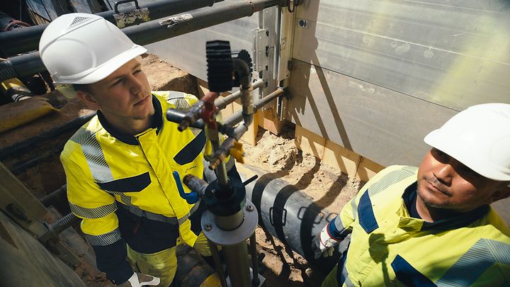 Zwei Personen mit Helmen arbeiten in einer Baugrube an Rohren und einem Ventilsystem.