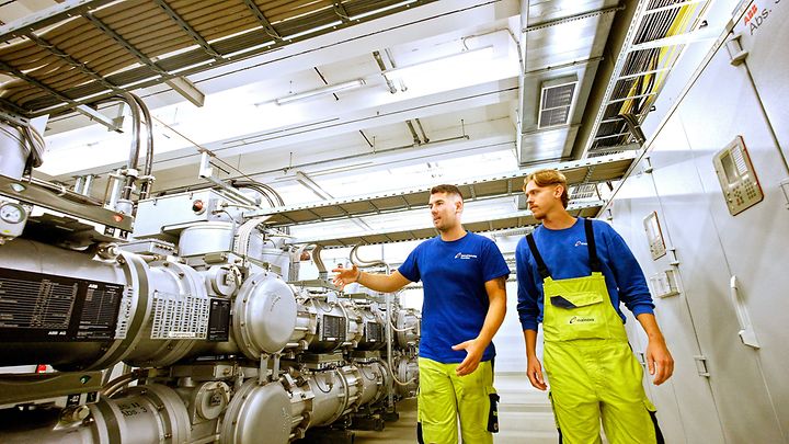 Zwei Personen mit Helmen arbeiten in einer Baugrube an Rohren und einem Ventilsystem.