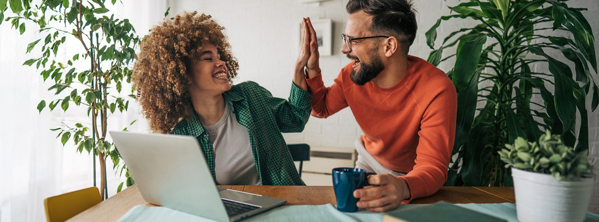 Zwei Personen sitzen am Tisch mit Laptop, lachen und geben sich einen High-Five beim Arbeiten.