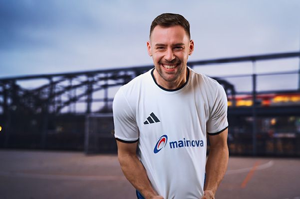 Person mit weißem Sporttrikot mit Mainova‑Logo steht im Vordergrund, im Hintergrund ein Sportplatz bei Dämmerung.