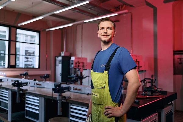 Man in einer Latzhose steht seitlich zur Kamera in einem Werkraum