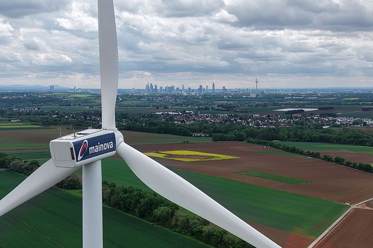 Windrad mit Mainova‑Logo über grünen Feldern, im Hintergrund die Frankfurter Skyline unter bewölktem Himmel.