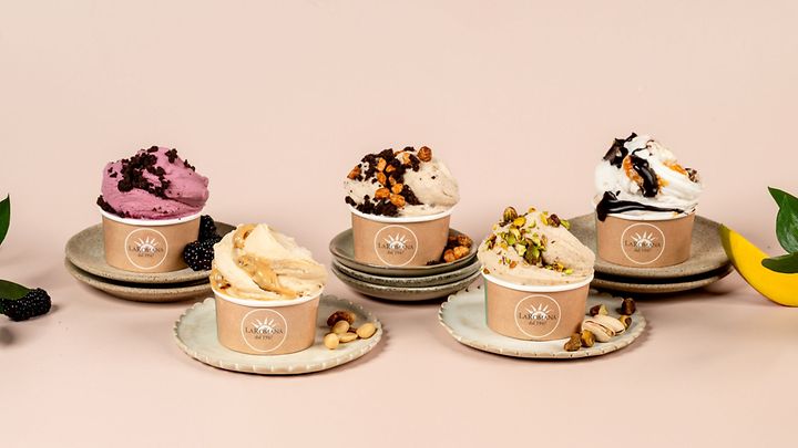 Fünf Becher Eis mit verschiedenen Toppings auf Tellern, umgeben von Nüssen, Früchten, vor hellem Hintergrund