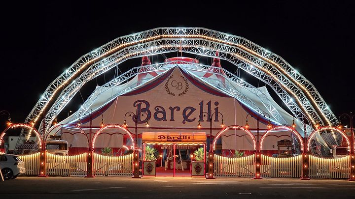Zirkuszelt „Barelli“ nachts, beleuchtet mit Lichterbögen, Eingangsbereich und nostalgischen Details im Vordergrund.