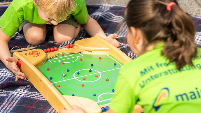 Junge und Mädchen in grünen Mainova T-Shirts spielen mit Tischfußball-Kasten auf einer Picknickdecke