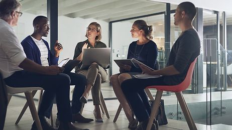 Fünf Personen mit Laptops sitzen in einem Stuhlkreis und unterhalten sich