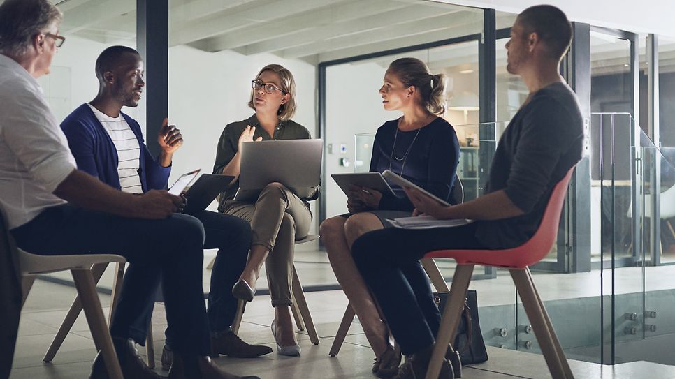 Fünf Personen mit Laptops sitzen in einem Stuhlkreis und unterhalten sich