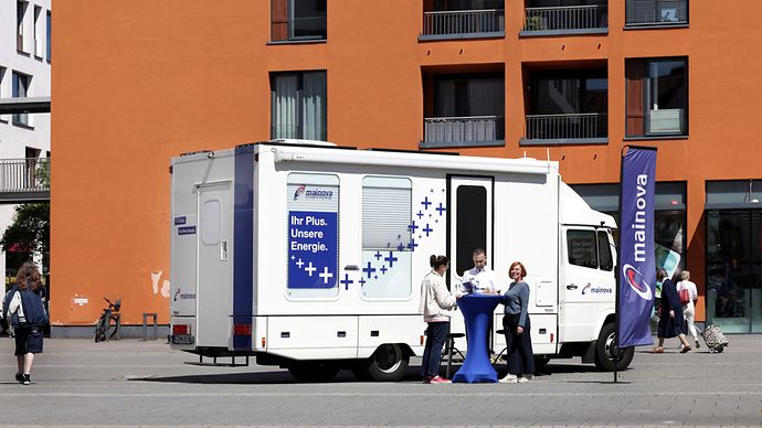 Infomobil der Mainova mit blauem Stehtisch, drei Personen im Gespräch und orangem Gebäude im Hintergrund.