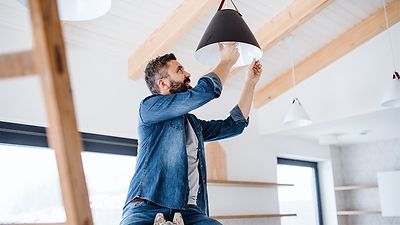 Mann sitzt auf einer Leiter und schraubt eine LED-Lampe in eine Deckenleuchte