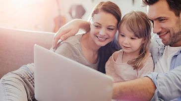 Frau und Mann mit kleiner Tochter sitzen eng zusammen auf dem Sofa und schauen auf einen Laptop