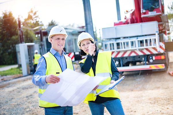 Mann und Frau mit Helm stehen mit Zeichnung am Baufahrzeug. Sie zeigt auf ein Gegenstand außerhalb des Bildes,