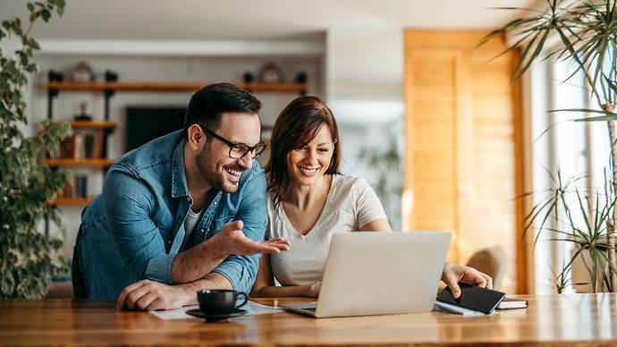 Paar sitzt an einem Holztisch und schaut gemeinsam auf einen Laptop, umgeben von Pflanzen und gemütlicher Einrichtung.