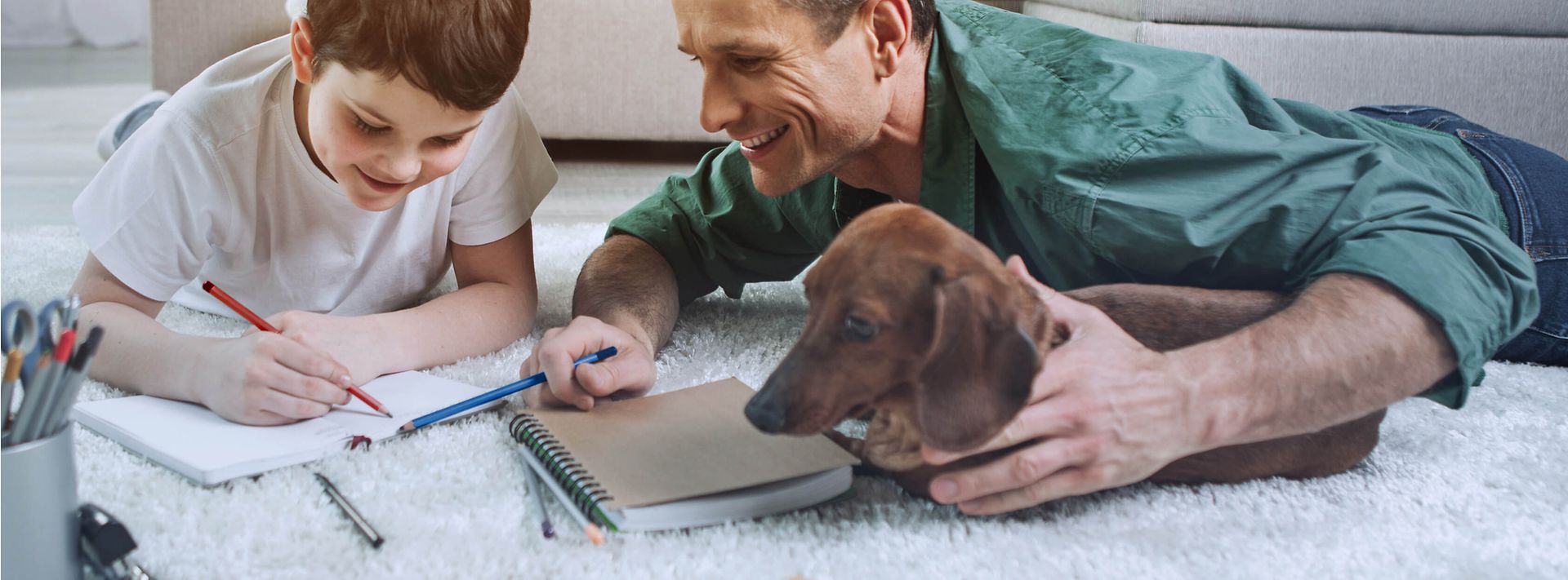 © iStock.com/YakobchukOlena Vater, Sohn und Dackel liegen auf dem Wohnzimmerteppich und machen schriftliche Hausaufgaben