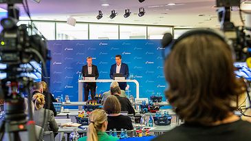 Konferenz mit zwei Sprechern auf der Bühne, umgeben von Kameras und Publikum in einem modernen Raum.