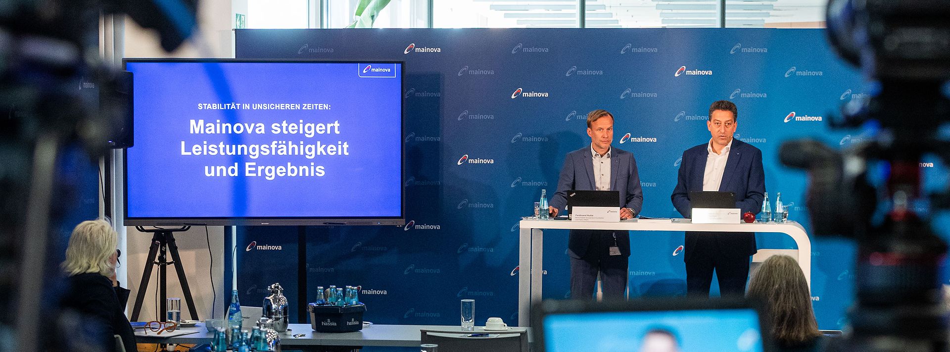 Zwei Personen bei Mainova-Pressekonferenz; Bildschirm mit Titel, Publikum sitzt, Kameras filmen.