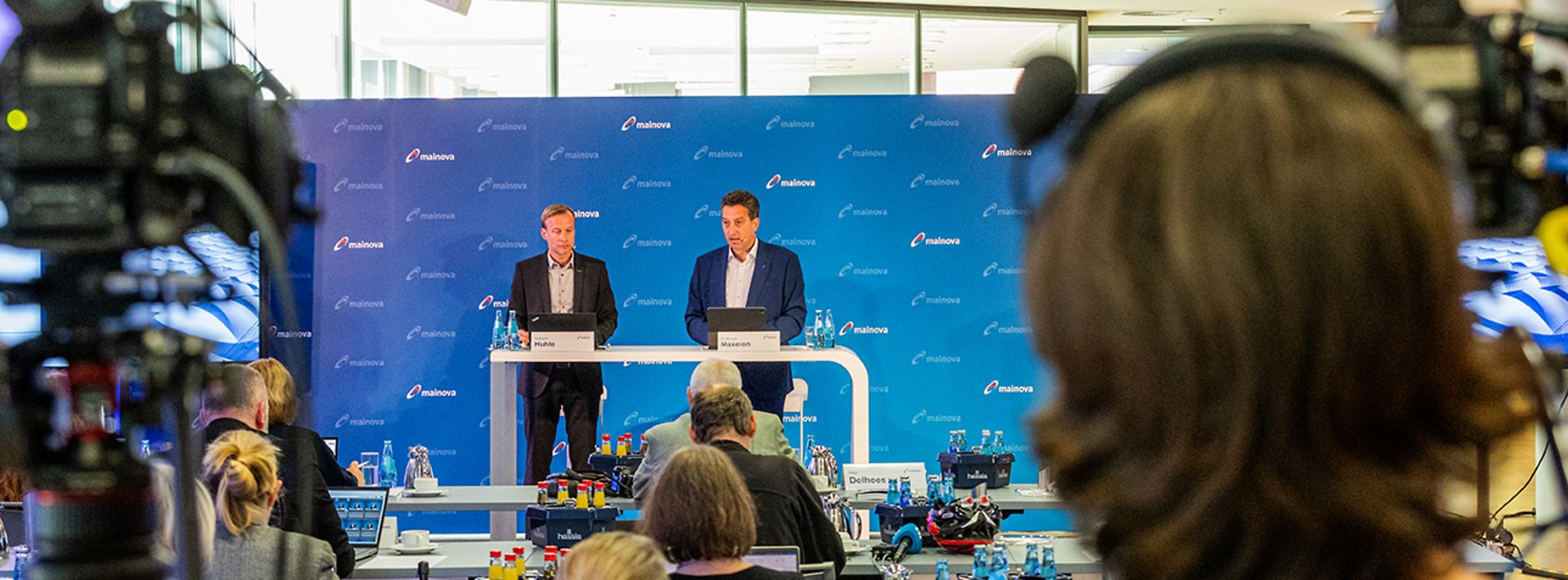 Konferenz mit zwei Sprechern auf der Bühne, umgeben von Kameras und Publikum in einem modernen Raum.