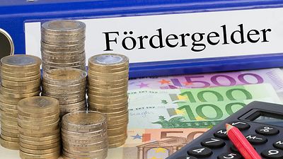 Ein Ordner mit ‚Fördergelder‘, gestapelte Münzen, Euro-Scheine, ein Taschenrechner und ein roter Stift