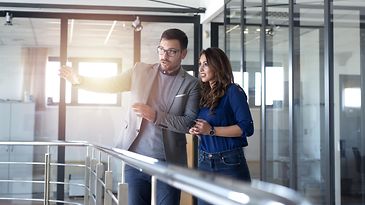 Ein Mann steht neben einer Arbeitskollegin im Büro und spricht mit ihr, während er in die Ferne zeigt.