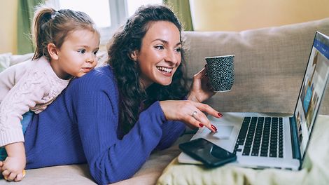 Frau liegt auf der Couch und surft am Laptop mit Tochter auf dem Rücken