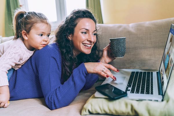 Frau liegt auf der Couch und surft am Laptop mit Tochter auf dem Rücken