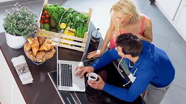 Eine Frau und ein Mann stehen an einem Tisch mit einem Laptop, einer Kiste Gemüse und Obst, Gebäck und einer Pflanze.