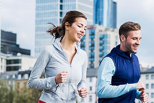 Zwei Personen joggen in Sportkleidung vor einer städtischen Kulisse mit modernen Gebäuden im Hintergrund.