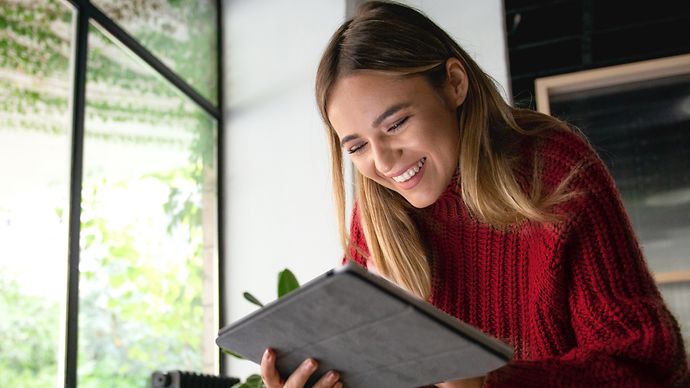 Eine Person in einem roten Pullover benutzt ein Tablet in einem Raum mit großen Fenstern und Pflanzen im Hintergrund.