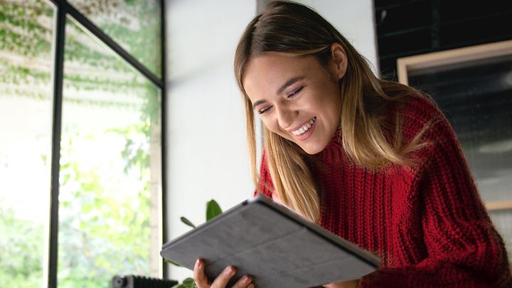 Eine Person in einem roten Pullover benutzt ein Tablet in einem Raum mit großen Fenstern und Pflanzen im Hintergrund.