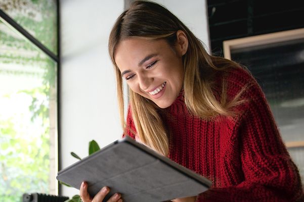 Eine Person in einem roten Pullover benutzt ein Tablet in einem Raum mit großen Fenstern und Pflanzen im Hintergrund.