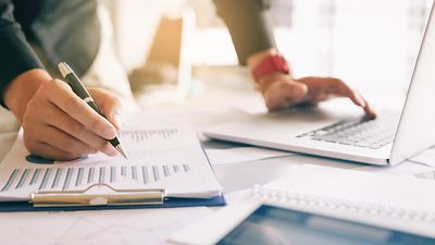 Eine Person schreibt auf ein Klemmbrett, während eine andere Hand einen Laptop bedient.