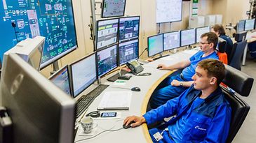 Zwei Personen in blauer Arbeitskleidung sitzen vor mehreren Computermonitoren in einem Kontrollraum.