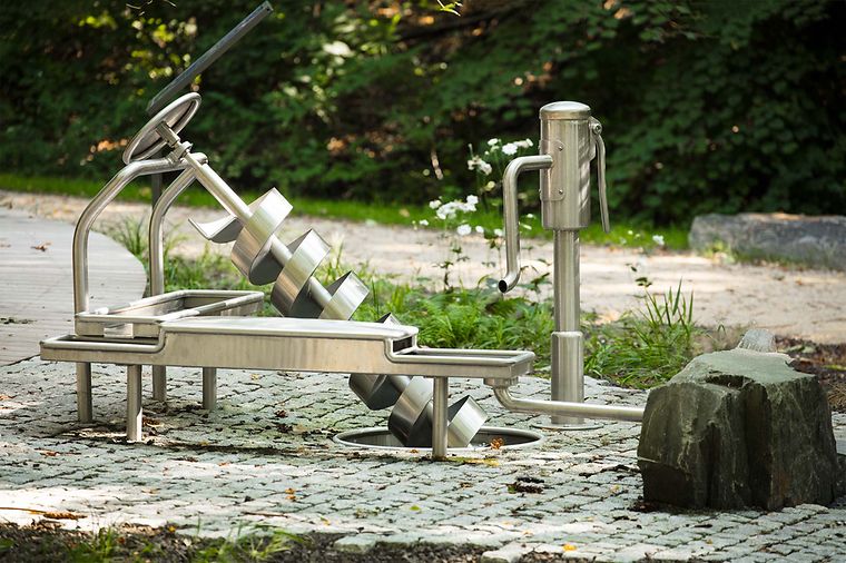 Ein Wasserspielplatz mit Edelstahlgeräten, darunter eine Wasserpumpe und schräg angeordnete Behälter.