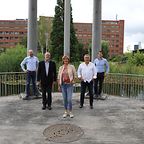 Fünf Personen stehen auf einer Holzplattform im Freien, umgeben von Säulen und einem Teich mit Schilf.