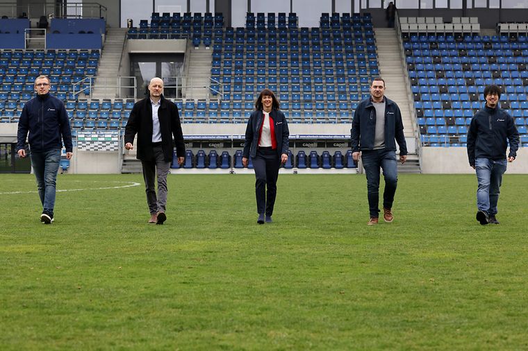 Fünf Personen gehen auf einem Fußballfeld in einem Stadion.