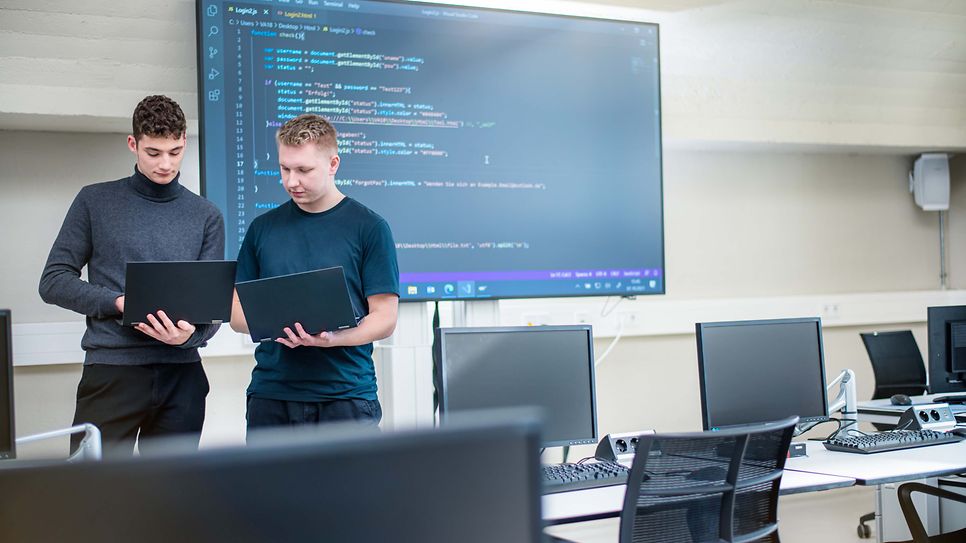 Zwei Auszubildende stehen zusammen und halten Notebooks in der Hand und vergleichen die Bildschirme.