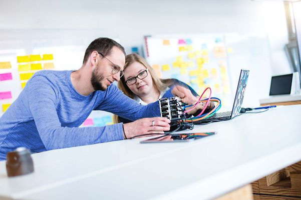 Ausbilder erklärt Auszubildende ein elektronisches Gerät was auf dem Tisch liegt