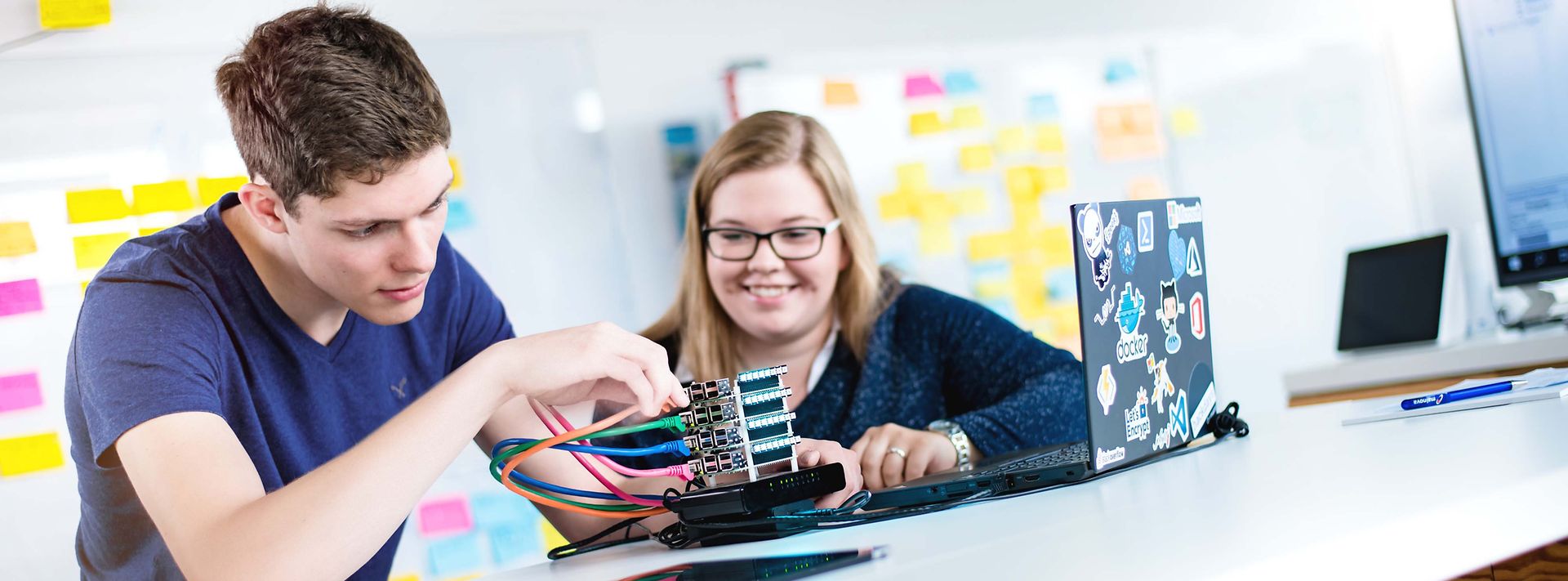 Zwei Auszubildende sitzen am Tisch und stecken Kabel in ein Elektronik Bauteil