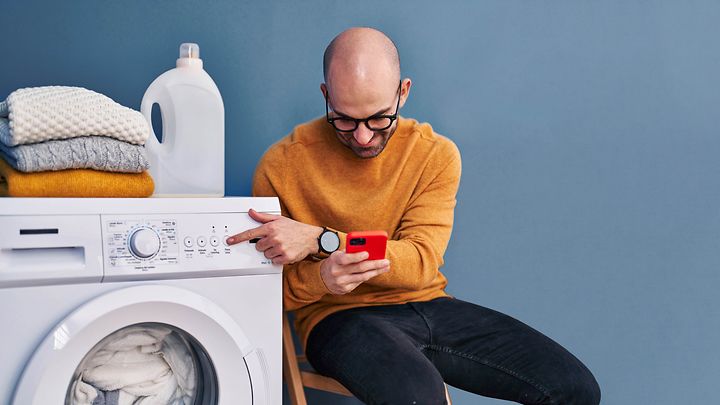 Mann mit Brille schaut auf sein Handy während er seine Waschmaschine bedient.