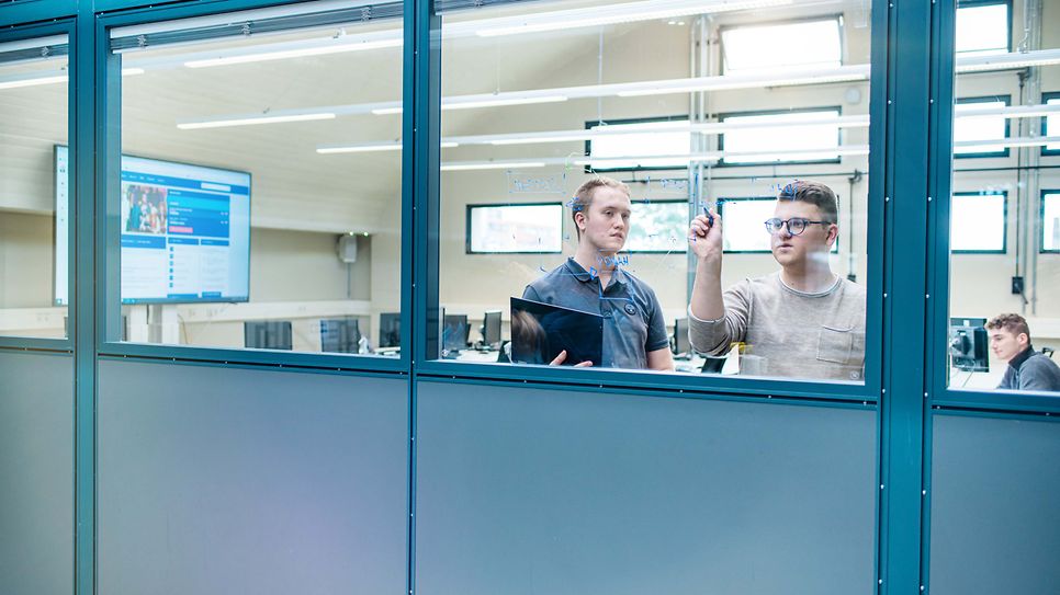 Zwei Auszubildende schreiben im Seminarraum etwas an die Scheibe. Fotografiert von Außen.